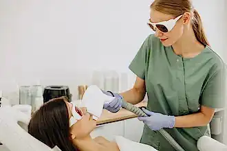 Gözlük takan bir estetisyen, bir kadına buz lazer epilasyon tedavisi uyguluyor.