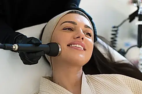 Baş bandı takan ve gülümseyen bir kadın, siyah eldiven giymiş bir estetisyenin Hydrafacial cilt bakım tedavisi uygulaması sırasında.