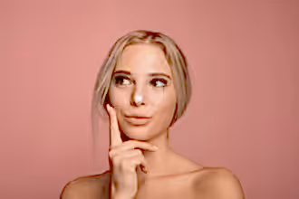 Pembe arka plan önünde, yüzüne krem süren ve gülen genç bir kadın.