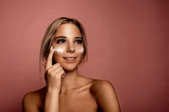 Pembe arka plan önünde, yüzüne krem süren genç bir kadın.