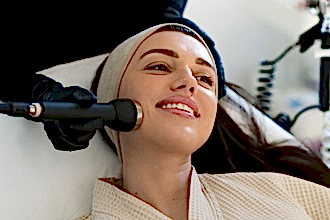 Siyah eldiven giymiş bir estetisyen, baş bandı takan ve gülümseyen bir kadının yüzüne Hydrafacial cihazıyla cilt bakım tedavisi uyguluyor.