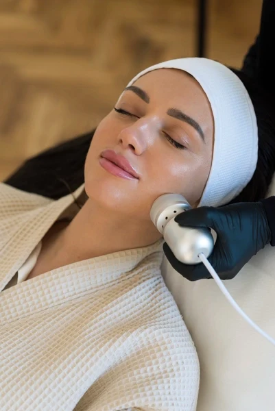 Siyah eldiven giymiş bir estetisyen, baş bandı takan bir kadına Hydrafacial cilt bakım tedavisi uyguluyor.