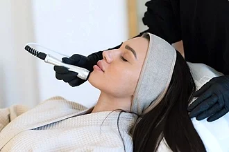Siyah eldiven giymiş bir estetisyen, baş bandı takan bir kadına Hydrafacial cilt bakım tedavisi uyguluyor.
