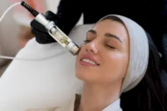 Siyah eldiven giymiş bir estetisyen, baş bandı takan ve gülümseyen bir kadına Hydrafacial cilt bakım tedavisi uyguluyor.